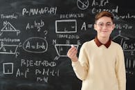 Young boy holding chalk in front of blackboard with math formulas, wearing glasses and smiling.
